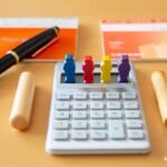 Calculadora com peças coloridas simbolizando pessoas, ao lado de itens bancários, representando organização financeira. Imagem LCbank.