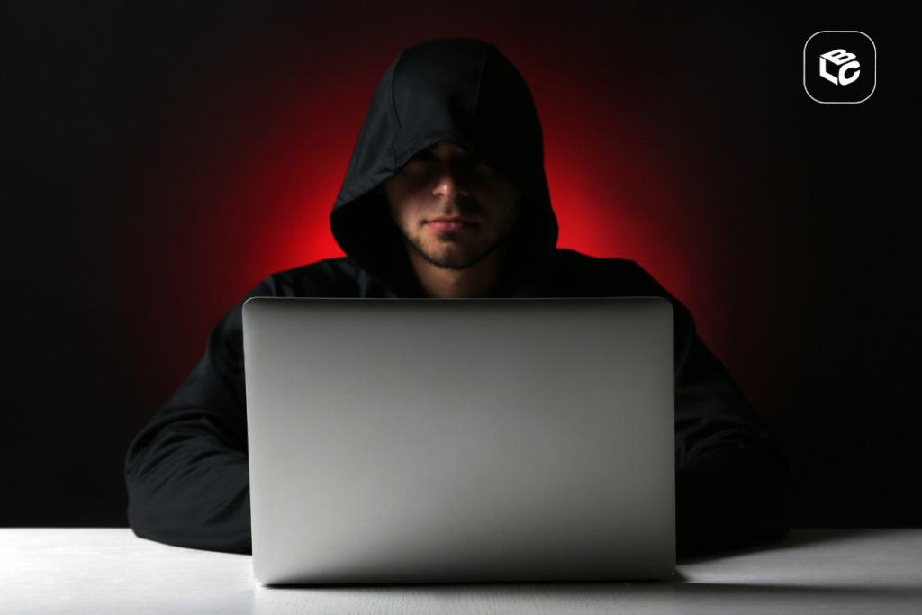 Pessoa usando moletom com capuz diante de um notebook em ambiente escuro, iluminado por luz vermelha ao fundo, representando segurança digital, cibercrime e ameaças virtuais