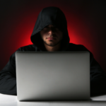 Pessoa usando moletom com capuz diante de um notebook em ambiente escuro, iluminado por luz vermelha ao fundo, representando segurança digital, cibercrime e ameaças virtuais